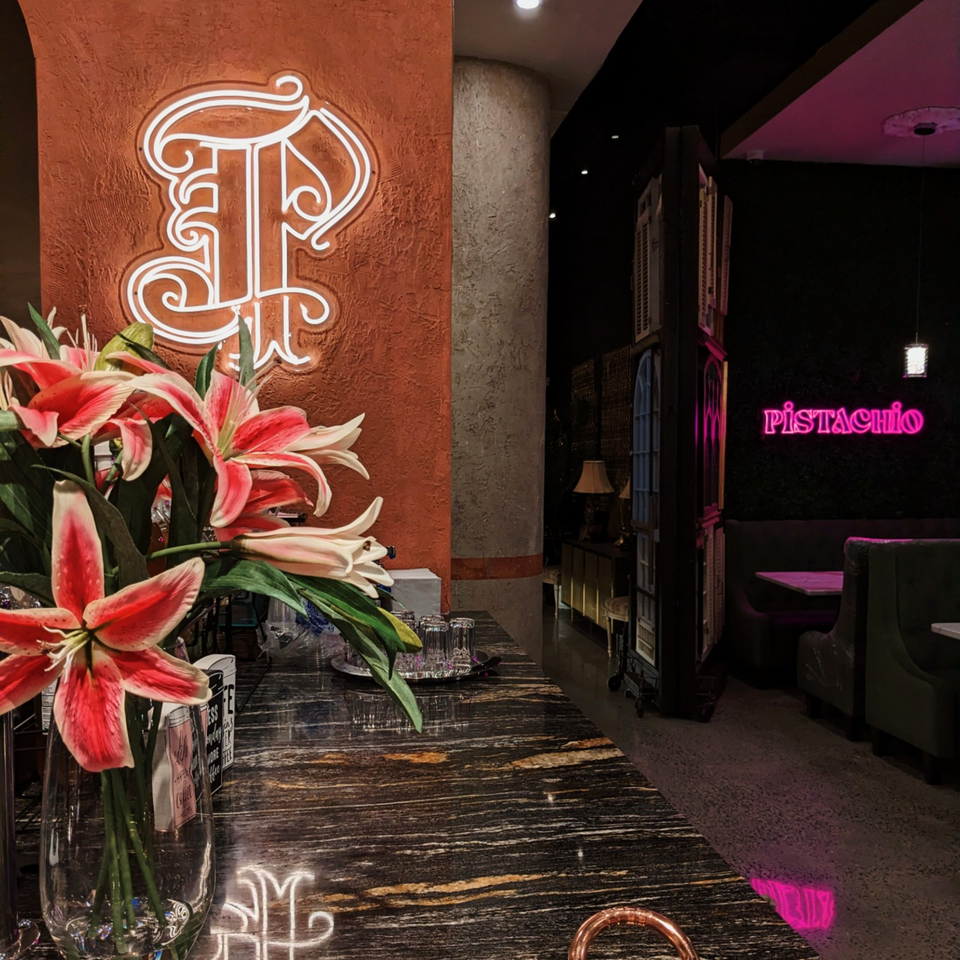 Restaurant entrance with pink lilies, ornate wall logo, dark dining booths, and a glowing Pistachio neon sign.