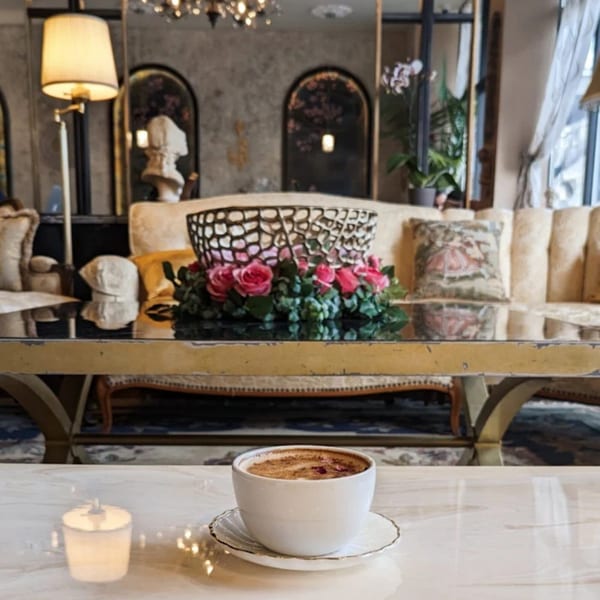 A cup of coffee on a marble table with a floral centerpiece and elegant cafe seating in the background.