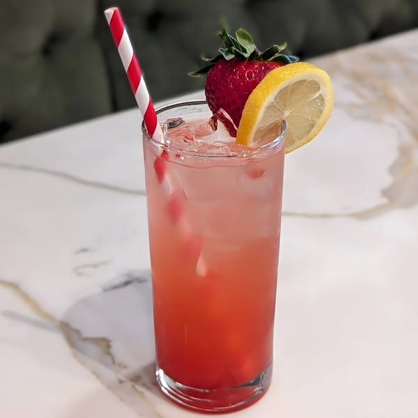 Pink drink with ice, a red and white straw, garnished with a strawberry and lemon slice.