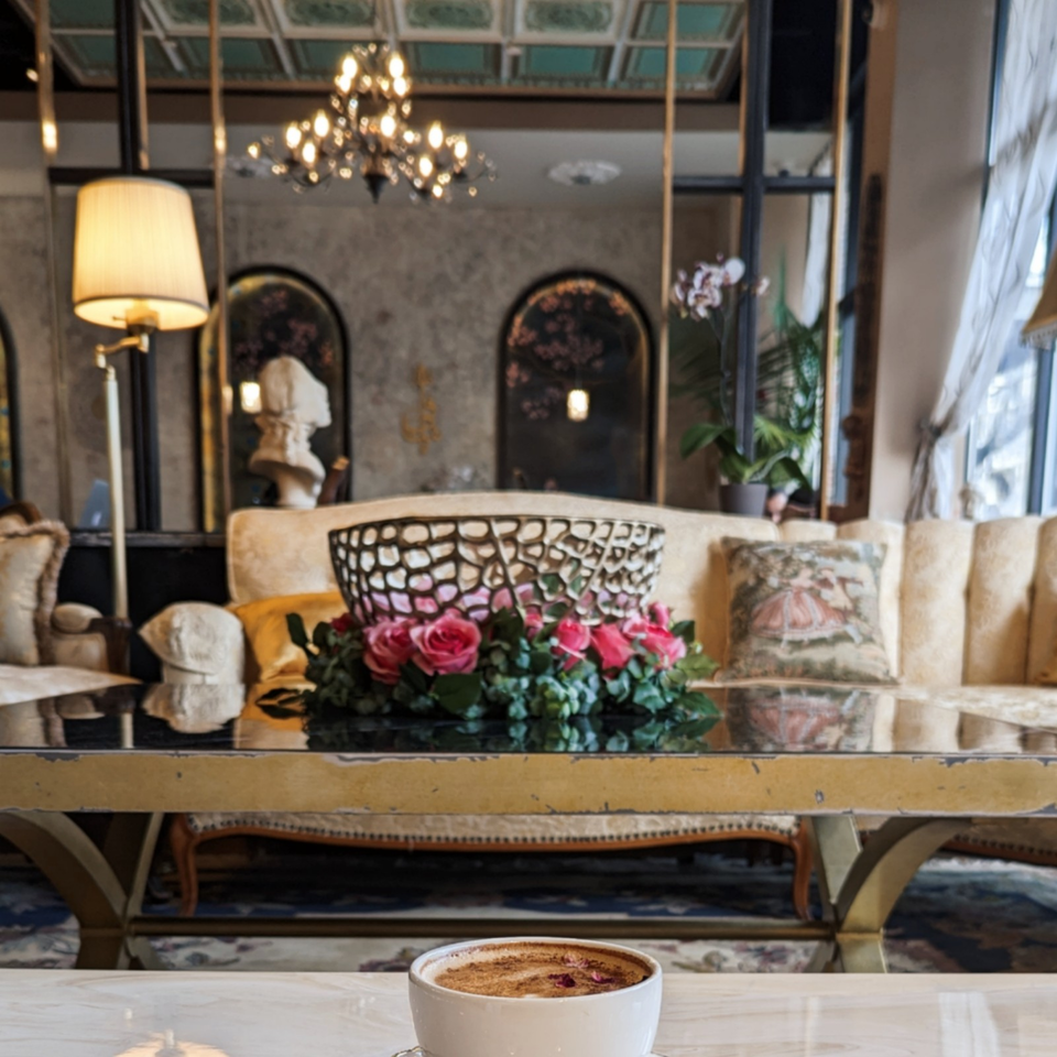 Coffee topped with foam and rose petals on a marble table in an elegant restaurant lounge with flowers, chandelier, and ornate furnishings.