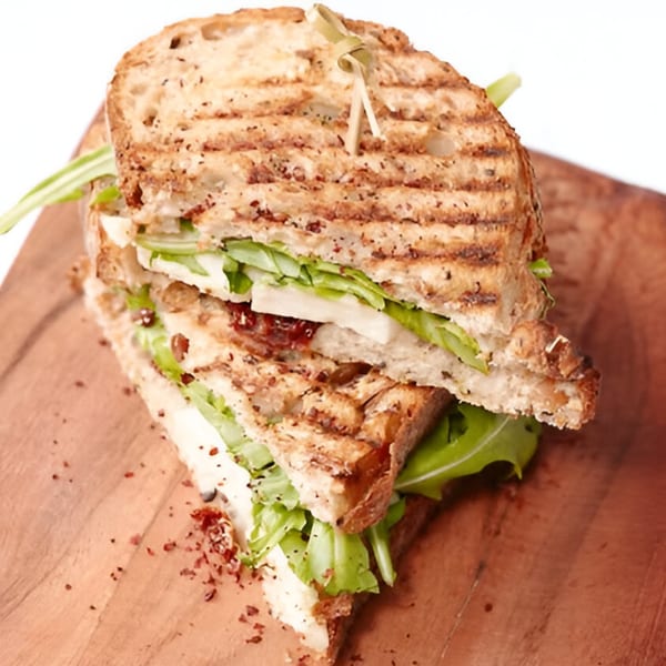 Grilled panini sandwich with cheese, greens, and a skewer, served on a wooden board at Pistachio Cafe.