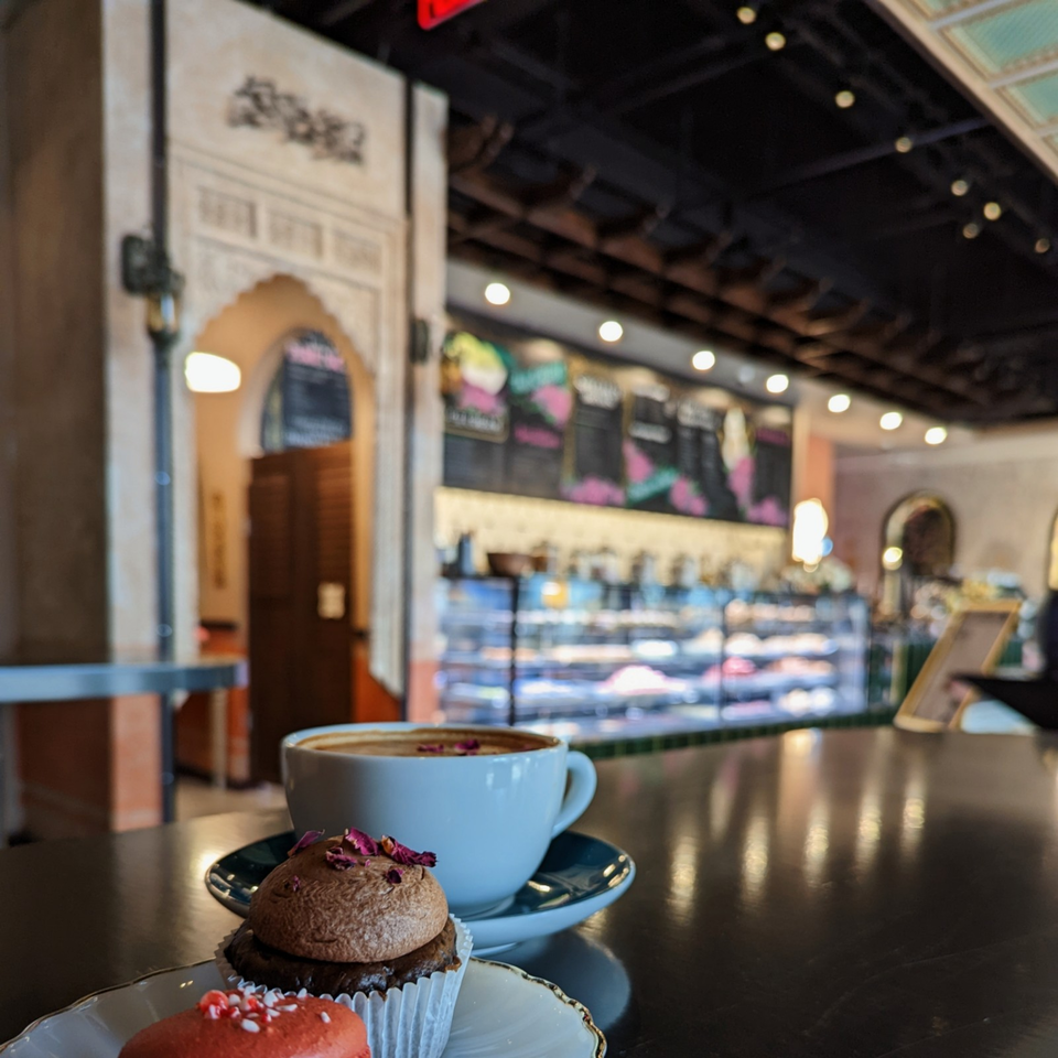 Coffee and decorated pastries sit on a café table overlooking a warmly lit bakery counter and chalkboard menu.