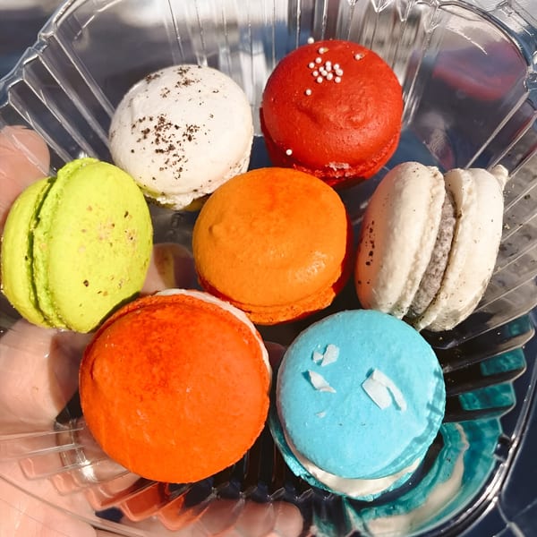 Seven colorful macarons in a plastic container, including red, orange, white, lime green, and blue.