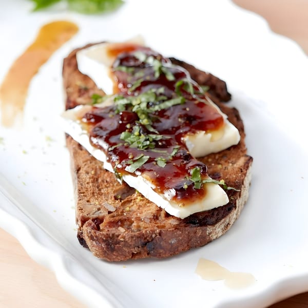 Toasted bread with melted brie cheese, dark jam, and fresh herbs on a white plate.