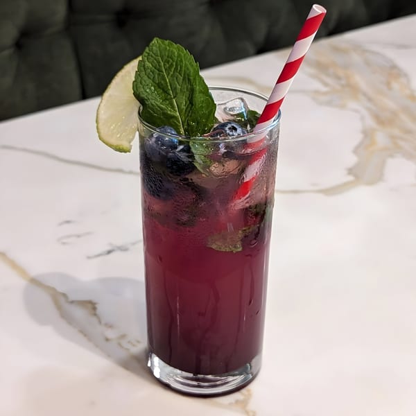 A tall glass of blueberry mojito with mint, lime, and a red-striped straw on a marble table.
