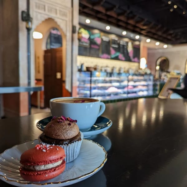 A red macaron, cupcake, and coffee on a table inside Pistachio Cafe, with display cases in the background.