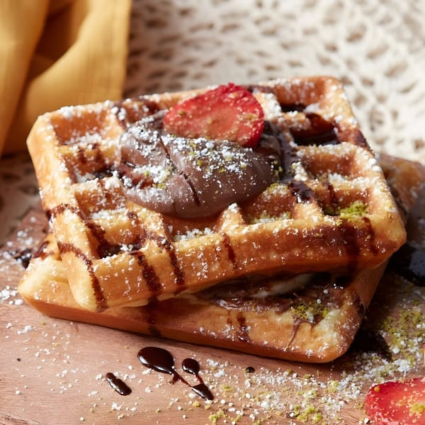 Two Pistachio Cafe waffles with chocolate, strawberries, powdered sugar, and pistachio crumbles.