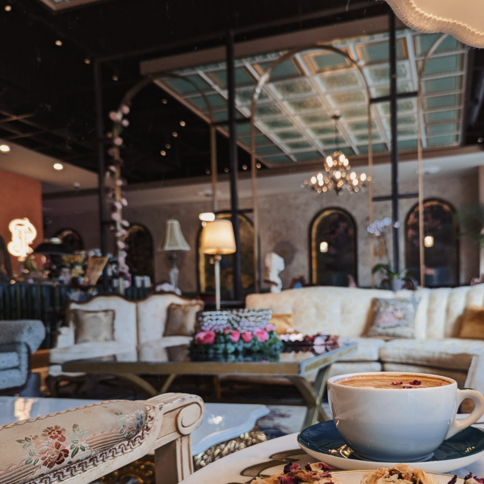 Cappuccino on a saucer beside a floral chair in an elegant café lounge with chandeliers, sofas, and a glass skylight.
