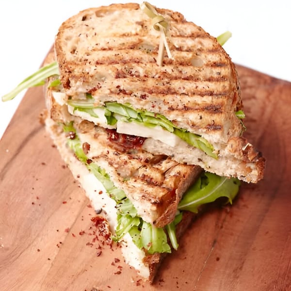 Grilled panini sandwich with cheese, arugula, and a dark spread on a wooden board.