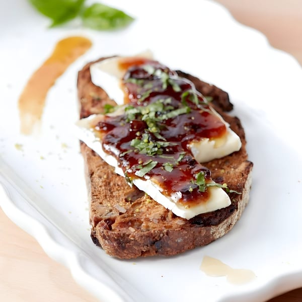 Toasted bread topped with brie cheese, dark jam, and fresh herbs on a white plate.