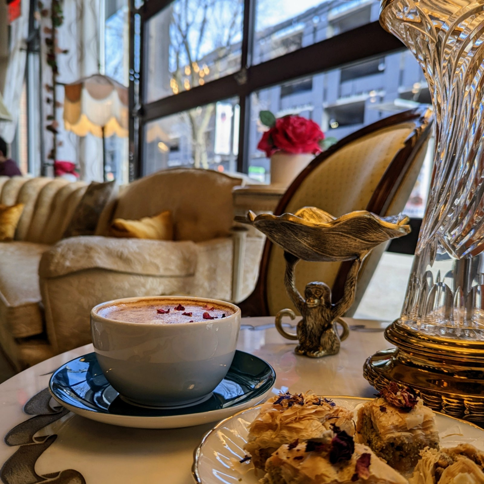 Cappuccino topped with rose petals and flaky pastries on a café table beside ornate décor and a velvet sofa.