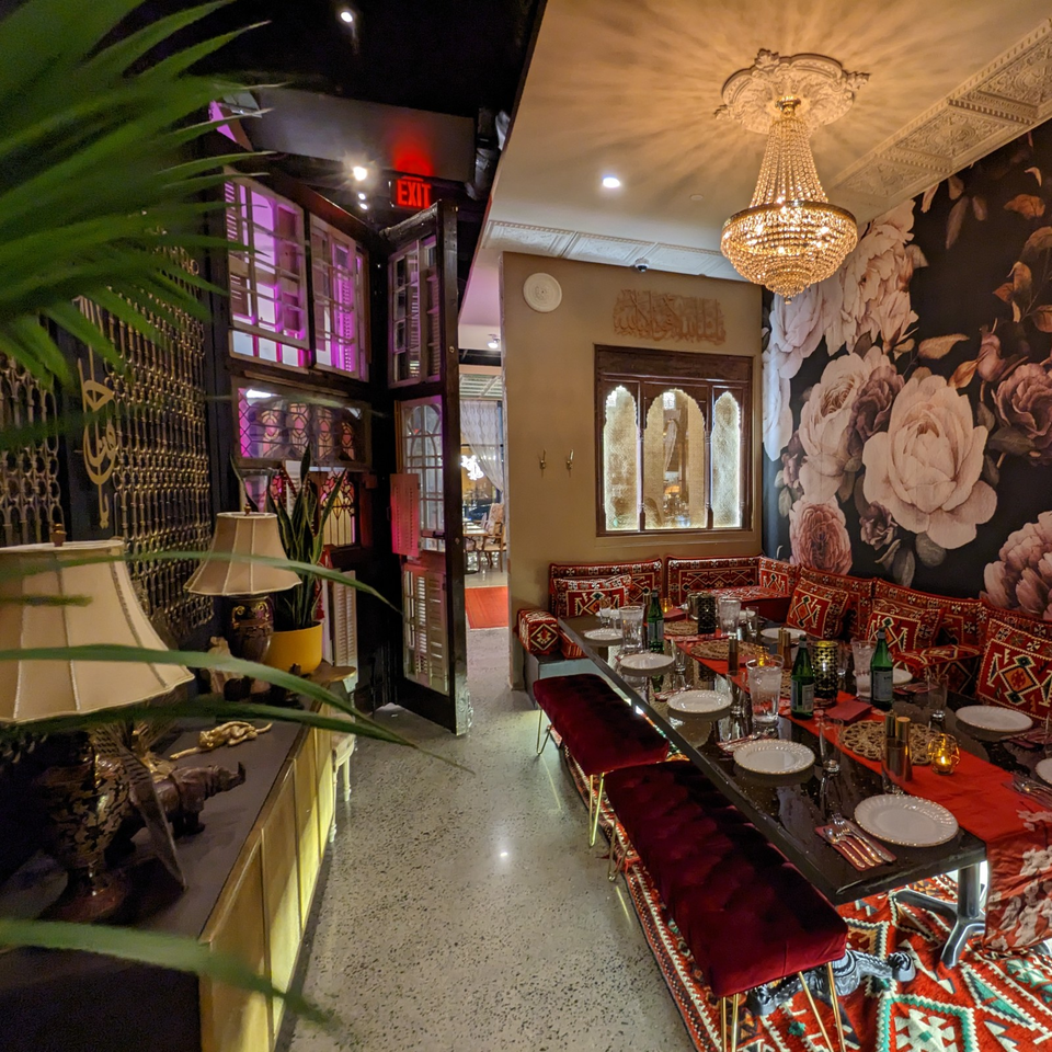 Ornate restaurant dining room with a crystal chandelier, floral mural, patterned cushions, red banquette seating, and set tables.