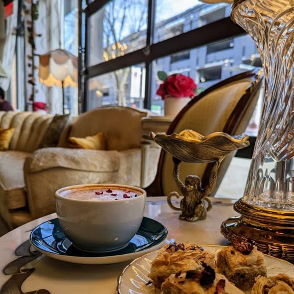 A latte with rose petals and baklava on a table at Pistachio Cafe, with a sofa and window in the background.