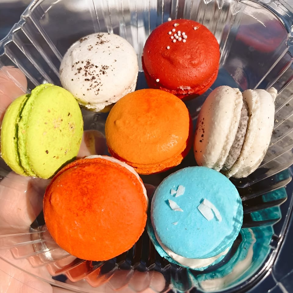 Seven colorful macarons in a clear plastic container, including red, orange, green, blue, and white.