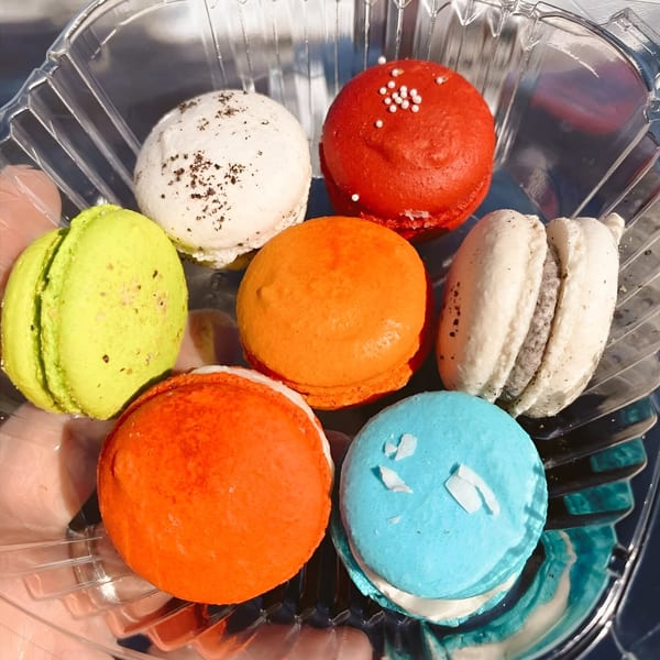 Seven colorful macarons in a clear plastic container, including red, orange, green, white, and blue.