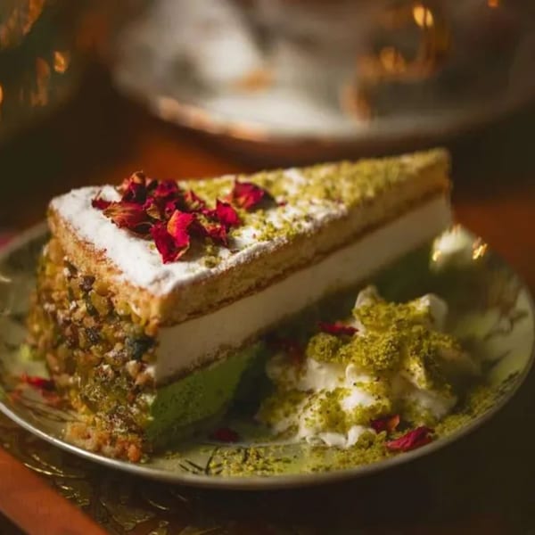 Slice of pistachio cake with rose petals, powdered sugar, and whipped cream on a plate.