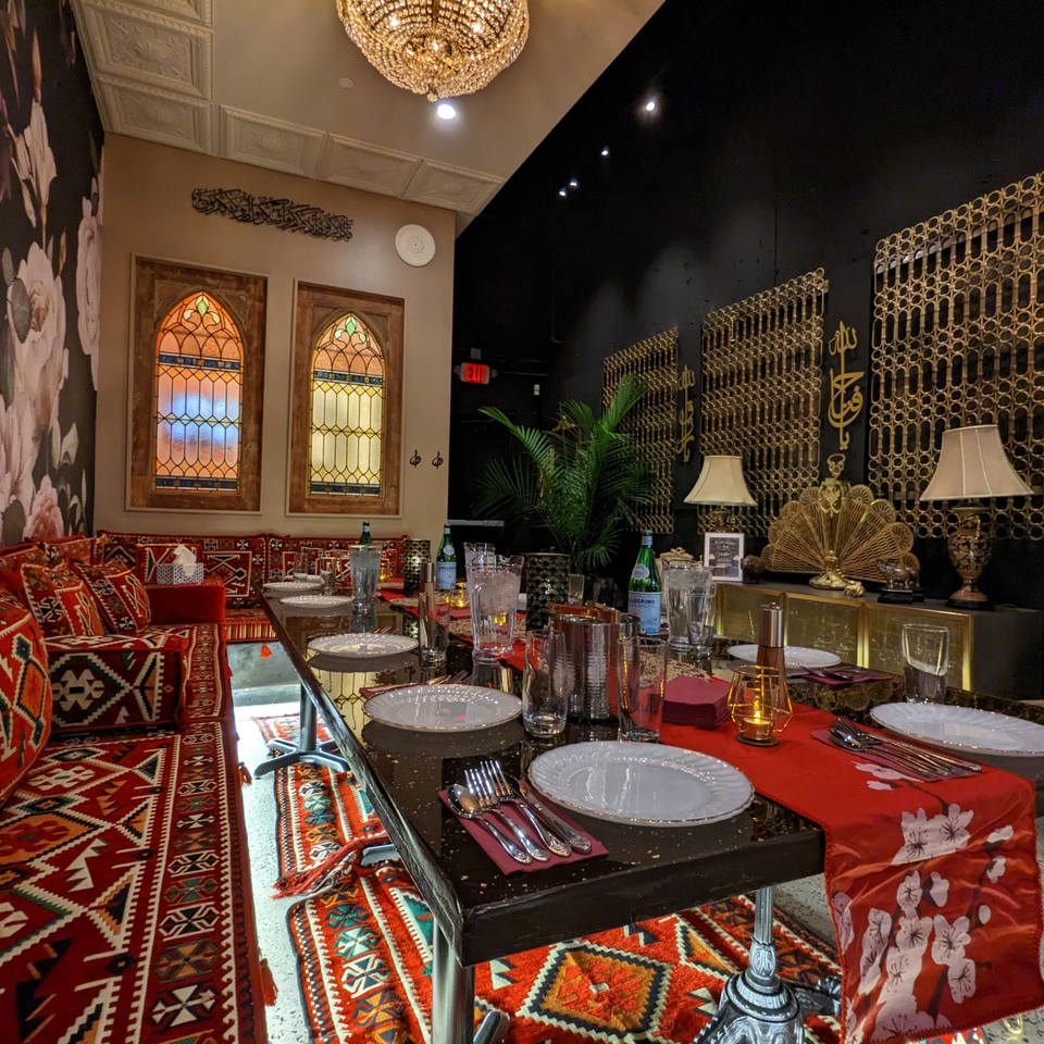 Luxurious restaurant dining room with ornate stained-glass windows, patterned red seating, gold decor, crystal chandelier, and fully set tables.