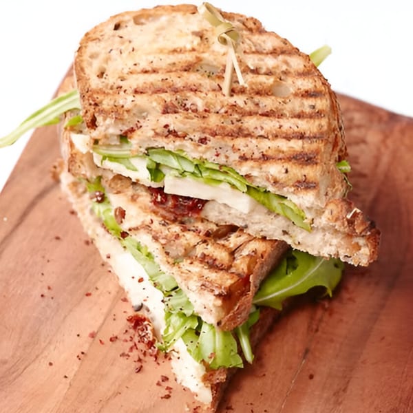 Grilled panini sandwich with cheese, lettuce, and sun-dried tomatoes on a wooden board.