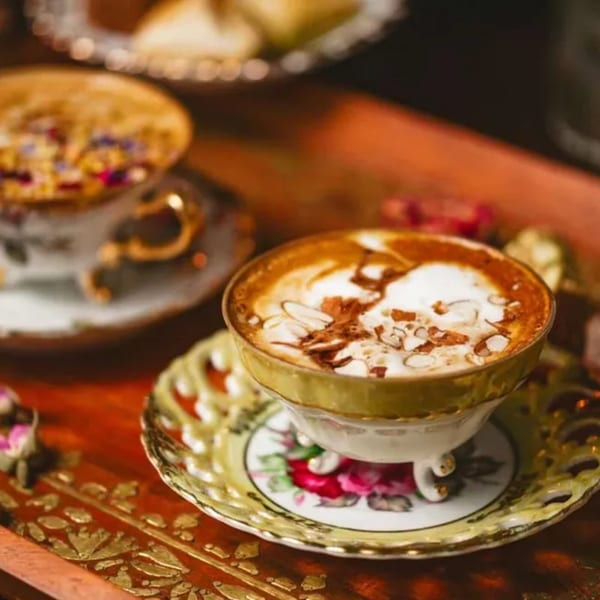 A pistachio latte with almond slivers and chocolate drizzle in an ornate gold and floral teacup.